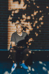 Lächelnder Teenager benutzt Smartphone, während er an einer Ziegelmauer sitzt - MASF20811