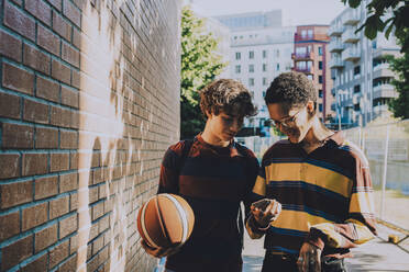 Smiling teenager holding soccer ball while male friend using phone by brick wall - MASF20816