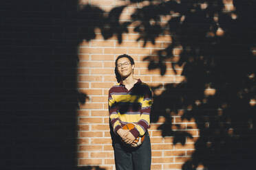 Smiling man with eyes closed holding soccer ball against brick wall - MASF20819