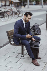 Entrepreneur with bag and file using smart phone while sitting on bench in city - MASF20830