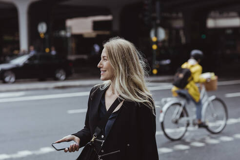 Lächelnde Geschäftsfrau mit Smartphone schaut in der Stadt weg - MASF20842