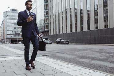 Männlicher Unternehmer mit Tasche, der beim Spaziergang in der Stadt ein Smartphone benutzt - MASF20848