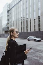 Side view of businesswoman with in-ear headphones holding file in city - MASF20858