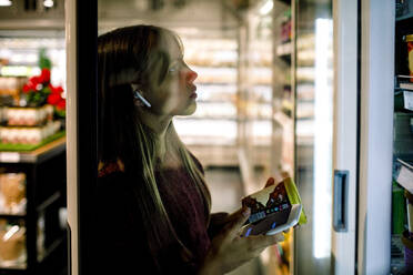 Weibliche Kundin mit In-Ohr-Kopfhörern und Mobiltelefon beim Einkaufen im Supermarkt - MASF20893
