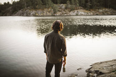 Rear view of young man standing by lake in forest during vacation - MASF20919