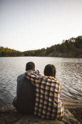 Rear view of man and woman sitting against lake in forest - MASF20937