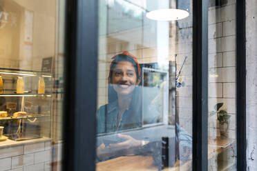 Junge Frau mit Mobiltelefon schaut durch das Fenster, während sie in einem Café sitzt - VABF03909