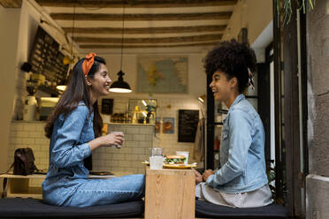 Smiling friends talking while sitting by window at cafe - VABF03918