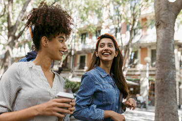 Woman holding coffee cup while walking with friend in city - VABF03938