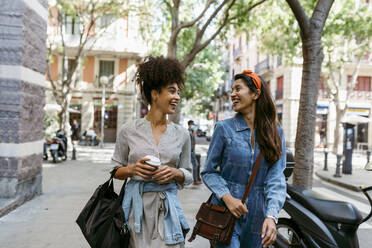 Smiling friends talking while walking at sidewalk in city - VABF03940