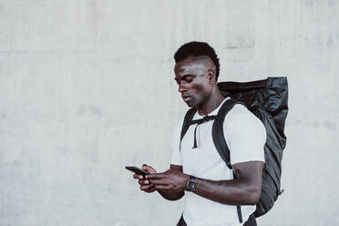 Sportler mit Rucksack Textnachrichten auf dem Smartphone, während er an der Wand steht - EBBF01343