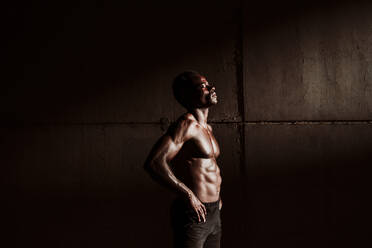 Nachdenklicher Sportler mit nackter Brust und Hand auf der Hüfte, der während des Sonnenuntergangs an einer grauen Wand steht - EBBF01367