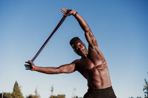 Male athlete doing resistance band exercise while standing against clear blue sky - EBBF01386