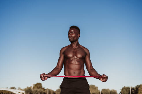 Male athlete looking away while stretching resistance band standing against clear sky during sunset - EBBF01389