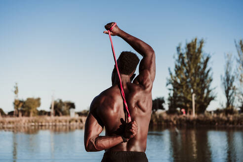 Young male athlete pulling resistance band while looking at sea against clear sky - EBBF01391