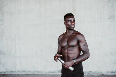 Male athlete looking away while holding water bottle standing against white wall - EBBF01394