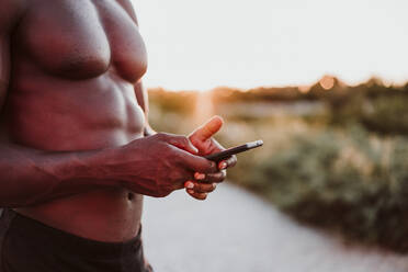 Männlicher Sportler, der sein Smartphone benutzt, während er bei Sonnenuntergang im Park steht - EBBF01405