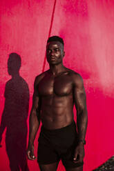 Male athlete looking away while standing by red wall during sunset - EBBF01407