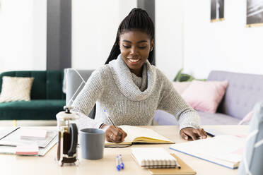 Smiling woman writing in book while sitting in living room at home - GIOF09573