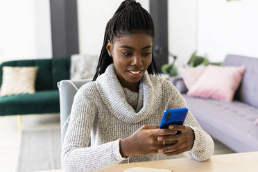 Smiling young woman text messaging on smart phone while sitting in living room at home - GIOF09576