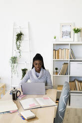 Female student concentrating while studying on laptop sitting at home - GIOF09587