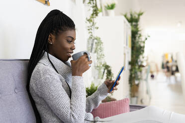 Junge Frau trinkt Kaffee, während sie zu Hause mit ihrem Smartphone SMS schreibt - GIOF09608