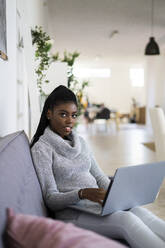 Junge Geschäftsfrau arbeitet am Laptop, während sie zu Hause auf dem Sofa sitzt - GIOF09620