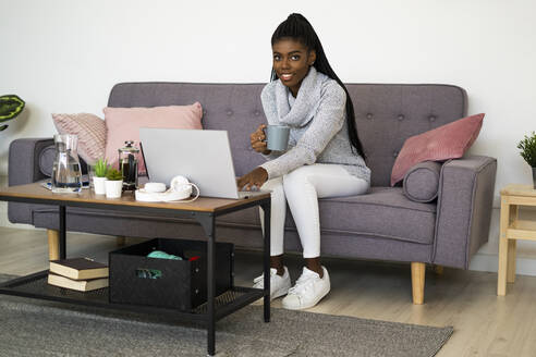Junge Geschäftsfrau trinkt Kaffee, während sie zu Hause am Laptop arbeitet - GIOF09629