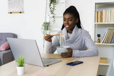 Woman eating noodles while watching movie on laptop sitting at home - GIOF09640