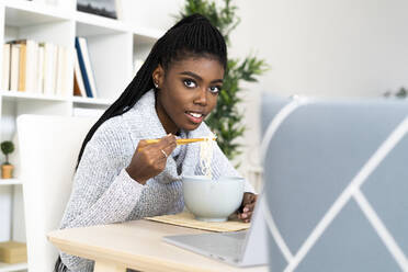 Junge Frau isst Nudeln mit Essstäbchen, während sie zu Hause sitzt - GIOF09644