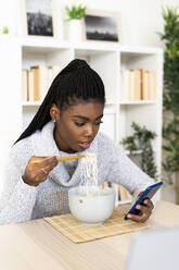 Young woman eating noodles text messaging on smart phone while sitting at home - GIOF09646