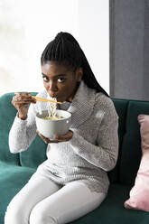 Young woman eating noodles while sitting on sofa at home - GIOF09647