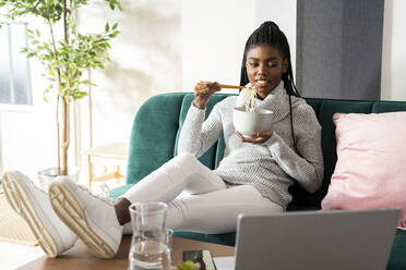 Young woman eating noodles while watching movie on laptop sitting in living room at home - GIOF09649