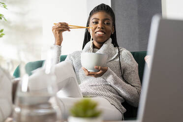 Smiling woman eating noodles while watching movie on laptop sitting in living room at home - GIOF09650