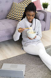 Young woman eating noodles while sitting on floor in living room at home - GIOF09651