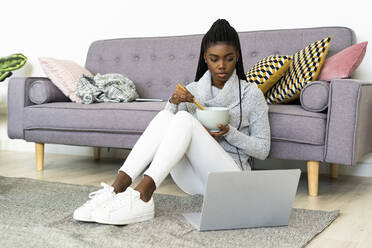 Junge Frau isst Nudeln, während sie zu Hause am Laptop einen Film ansieht - GIOF09653