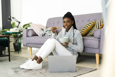 Smiling woman eating noodles while sitting on floor in living room at home - GIOF09654