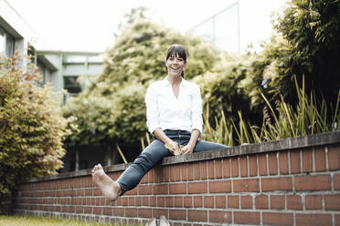 Smiling female professional sitting on retaining wall by plants at industry - JOSEF02141