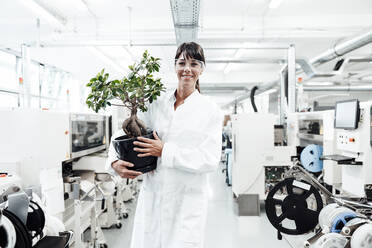 Smiling female scientist holding potted plant while standing amidst machineries at laboratory - JOSEF02177
