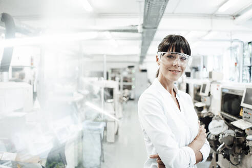 Smiling female engineer wearing protective eyewear while standing with arms crossed in industry - JOSEF02202