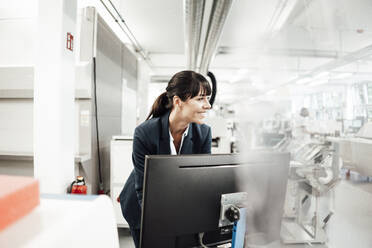 Smiling businesswoman looking away while standing in front of computer monitor at industry - JOSEF02203