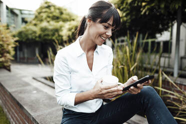 Smiling female professional holding coffee cup while using smart phone during break - JOSEF02235