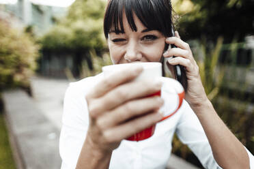 Lächelnde Geschäftsfrau, die eine Kaffeetasse hält, während sie mit einem Mobiltelefon spricht - JOSEF02236