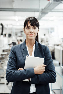 Confident businesswoman holding digital tablet while standing with arms crossed at industry - JOSEF02258