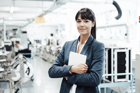 Smiling businesswoman holding digital tablet while standing with arms crossed at industry - JOSEF02259