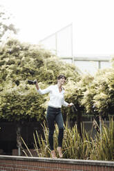 Smiling carefree businesswoman holding shoe walking on retaining wall at industry - JOSEF02294