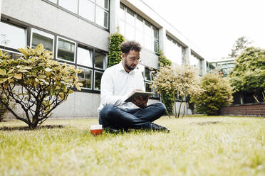 Businessman using digital tablet while sitting with coffee cup on grass against industry - JOSEF02295