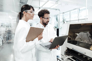 Male and female scientists with laptop and clipboard looking at machinery in laboratory - JOSEF02297