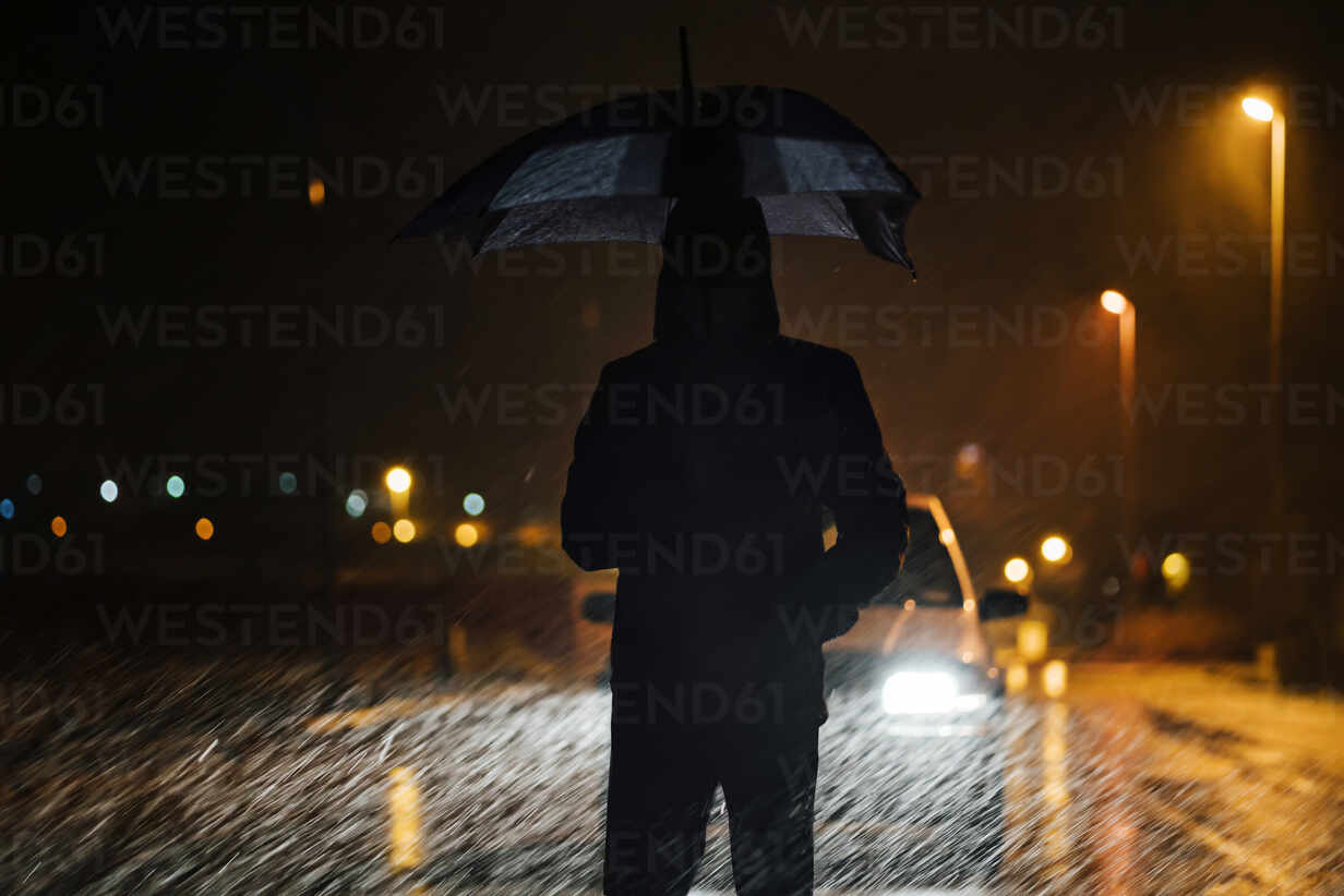 Junger Mann mit Regenschirm steht in einer regnerischen Nacht vor