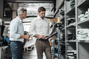 Businessmen discussing over equipment while standing by rack in factory - JOSEF02309